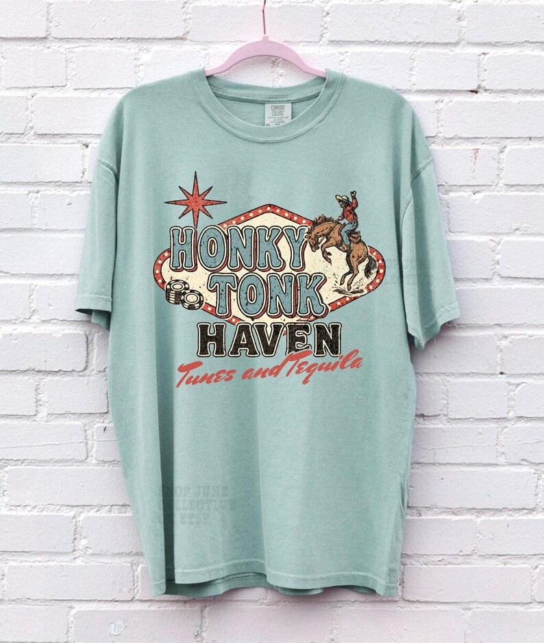 Pode incluir: Uma camiseta verde claro com um design gr&aacute;fico vintage que diz "Honky Tonk Haven". O design inclui um cowboy montando um cavalo e uma estrela vermelha. O texto "Tunes and Tequila" tamb&eacute;m est&aacute; inclu&iacute;do.