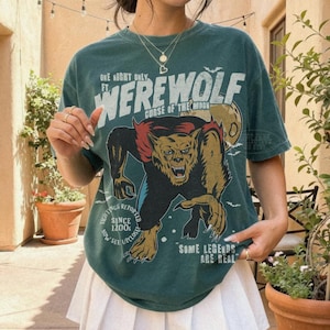 May include: A dark green t-shirt featuring a graphic of a werewolf with the text "ONE NIGHT ONLY FT. WEREWOLF CURSE OF THE MOON", "SIGHTINGS REPORTED SINCE 1200S NOW SEE UPCLOSE", and "SOME LEGENDS ARE REAL" paired with a white pleated mini skirt.