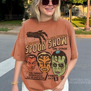 May include: A rust-colored short-sleeve t-shirt featuring a graphic print with a clawed hand, the text "MIDNIGHT SPOOK SHOW", "FT. REAL MONSTERS", "ON STAGE", "THE CREEPIEST HORROR SHOW YOU'VE EVER SEEN", and "FRIDAY OCT. 13" above three monster faces.