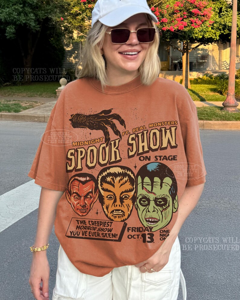 May include: A rust-colored short-sleeve t-shirt featuring a graphic print with a clawed hand, the text "MIDNIGHT SPOOK SHOW", "FT. REAL MONSTERS", "ON STAGE", "THE CREEPIEST HORROR SHOW YOU'VE EVER SEEN", and "FRIDAY OCT. 13" above three monster faces.