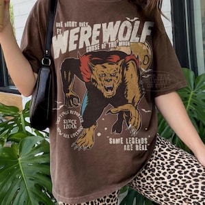 May include: A brown short-sleeve t-shirt featuring a graphic of a werewolf with the text "ONE NIGHT ONLY FT. WEREWOLF CURSE OF THE MOON", "SIGHTINGS REPORTED SINCE 1200s NOW SEE UPCLOSE", and "SOME LEGENDS ARE REAL" printed in cream and black.