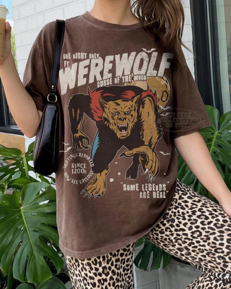 May include: A brown short-sleeve t-shirt featuring a graphic of a werewolf with the text "ONE NIGHT ONLY FT. WEREWOLF CURSE OF THE MOON", "SIGHTINGS REPORTED SINCE 1200s NOW SEE UPCLOSE", and "SOME LEGENDS ARE REAL" printed in cream and black.