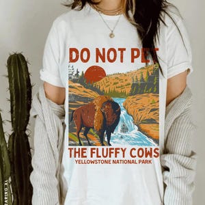 May include: White t-shirt featuring a vintage-style graphic of a buffalo in a mountain landscape. The text "Do Not Pet The Fluffy Cows Yellowstone National Park" is printed on the shirt.