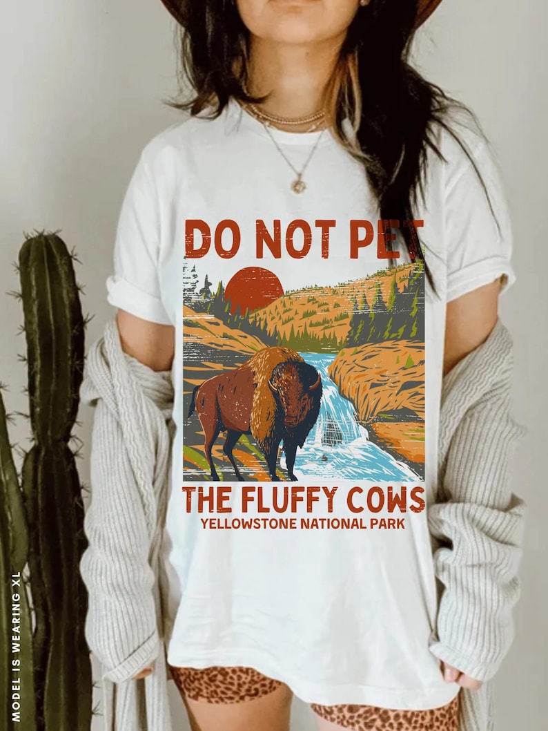 May include: White t-shirt featuring a vintage-style graphic of a buffalo in a mountain landscape. The text "Do Not Pet The Fluffy Cows Yellowstone National Park" is printed on the shirt.
