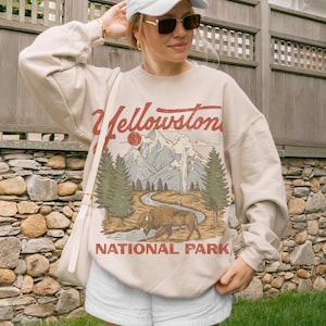 May include: A beige crewneck sweatshirt featuring a graphic print of Yellowstone National Park with a bison, pine trees, mountains, and a geyser. The text Yellowstone National Park is printed in red and brown lettering across the front of the garment.