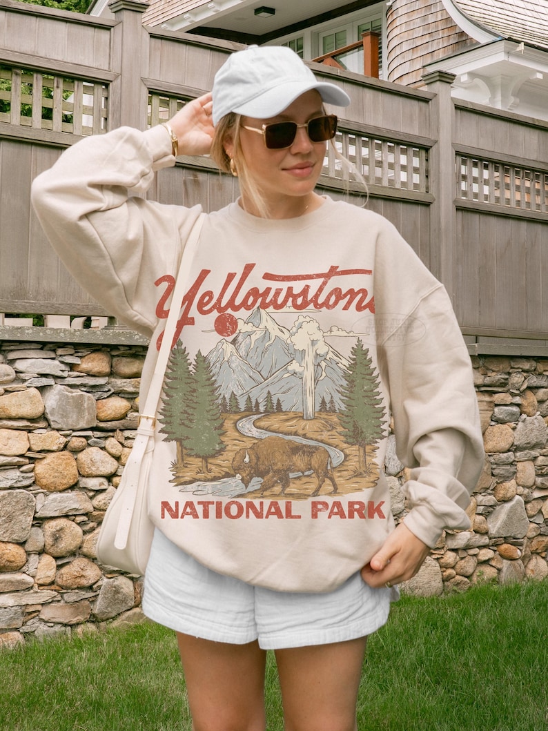 May include: A beige crewneck sweatshirt featuring a graphic print of Yellowstone National Park with a bison, pine trees, mountains, and a geyser. The text Yellowstone National Park is printed in red and brown lettering across the front of the garment.