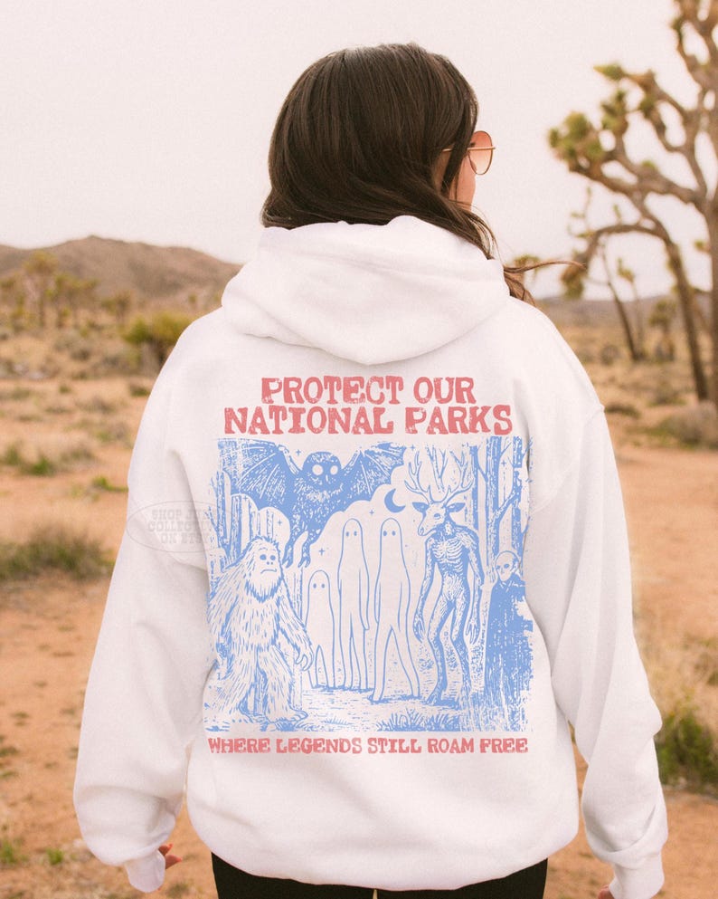 May include: A white hooded sweatshirt featuring a blue graphic print on the back. The design depicts a forest scene with a large owl, a sasquatch, two ghosts, a deer-like creature, and a figure. Text reads: PROTECT OUR NATIONAL PARKS WHERE LEGENDS STILL ROAM FREE.