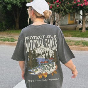 May include: A gray t-shirt with a graphic print of a mountain scene and a fish. The text "PROTECT OUR NATIONAL PARKS" is printed on the shirt. The text "Explore Responsibly" is printed below the graphic.