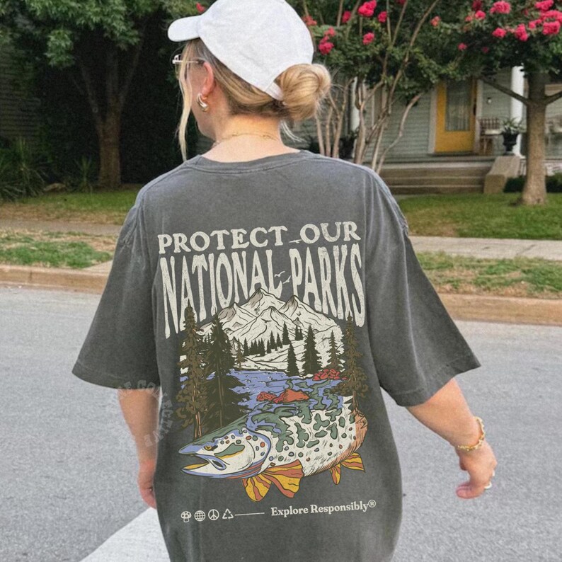 May include: A gray t-shirt with a graphic print of a mountain scene and a fish. The text "PROTECT OUR NATIONAL PARKS" is printed on the shirt. The text "Explore Responsibly" is printed below the graphic.