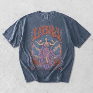 May include: A charcoal gray short-sleeve t-shirt featuring a graphic print of a person holding scales. The text LIBRA is arched above the figure, with orange, pink, and blue colors in the illustration, including floral motifs and a zodiac symbol.