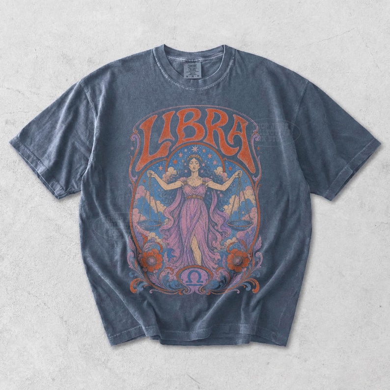 May include: A charcoal gray short-sleeve t-shirt featuring a graphic print of a person holding scales. The text LIBRA is arched above the figure, with orange, pink, and blue colors in the illustration, including floral motifs and a zodiac symbol.