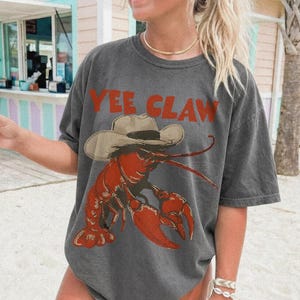 Op de afbeelding: Donkergrijs t-shirt met een rode kreeft met een cowboyhoed. De tekst "Yee Claw" staat boven de kreeft. Casual stijl.