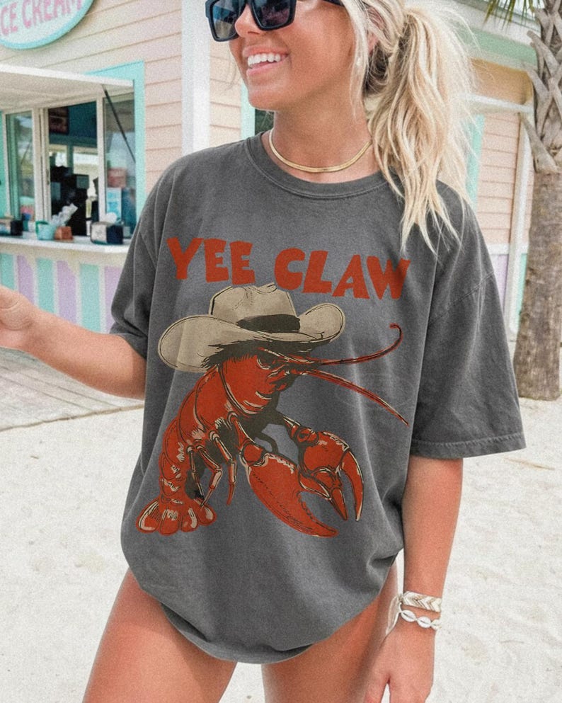Op de afbeelding: Donkergrijs t-shirt met een rode kreeft met een cowboyhoed. De tekst "Yee Claw" staat boven de kreeft. Casual stijl.