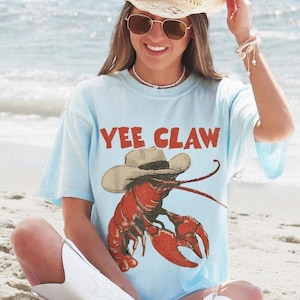 Op de afbeelding: Lichtblauw T-shirt met de rode tekst "YEE CLAW", met een cartoon kreeft met een cowboyhoed. De persoon draagt een strooien cowboyhoed en witte cowboylaarzen.