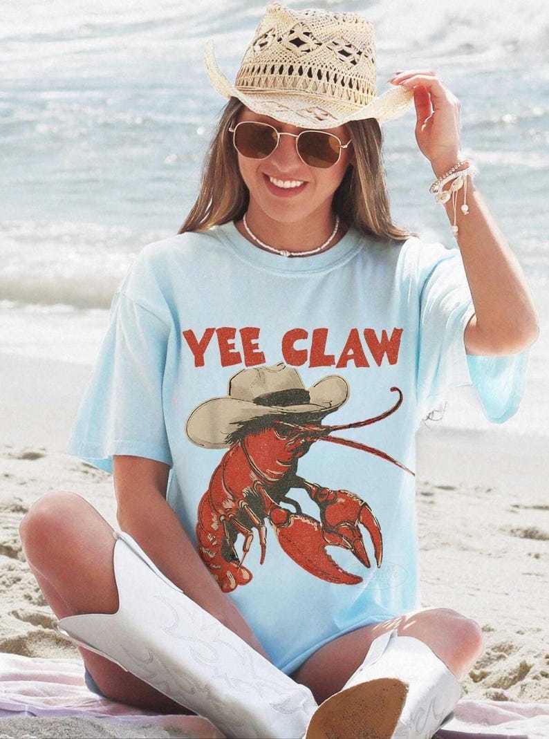 Op de afbeelding: Lichtblauw T-shirt met de rode tekst "YEE CLAW", met een cartoon kreeft met een cowboyhoed. De persoon draagt een strooien cowboyhoed en witte cowboylaarzen.