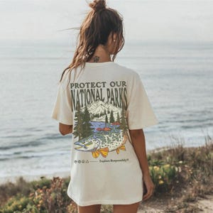 May include: A white t-shirt with a graphic print of a mountain scene and a fish. The text "Protect Our National Parks" is printed on the shirt. The text "Explore Responsibly" is printed below the graphic.