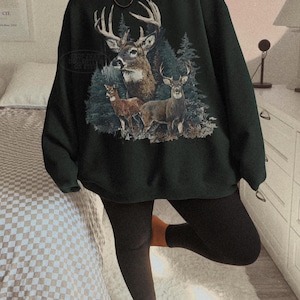 Puede incluir: Sudadera verde oscuro con un gr&aacute;fico de tres ciervos en un bosque. El dise&ntilde;o muestra un gran ciervo con cornamenta y dos ciervos m&aacute;s peque&ntilde;os. La sudadera es de estilo casual.