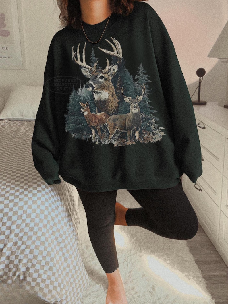 Puede incluir: Sudadera verde oscuro con un gr&aacute;fico de tres ciervos en un bosque. El dise&ntilde;o muestra un gran ciervo con cornamenta y dos ciervos m&aacute;s peque&ntilde;os. La sudadera es de estilo casual.