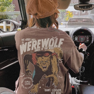 May include: A brown t-shirt featuring a graphic print of a werewolf with the text "ONE NIGHT ONLY FT. WEREWOLF CURSE OF THE MOON" and "SOME LEGENDS ARE REAL" along with "SIGHTINGS REPORTED SINCE 1200S NOW SEE UPCLOSE". The shirt is worn with a brown corduroy cap.