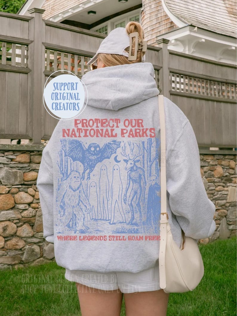 May include: A light grey hooded sweatshirt featuring a blue and red graphic on the back. The text reads "PROTECT OUR NATIONAL PARKS" and "WHERE LEGENDS STILL ROAM FREE" with illustrations of a sasquatch, an owl, ghosts, and a deer-like creature.