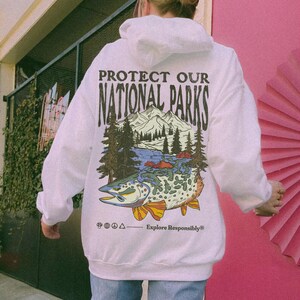May include: A white hooded sweatshirt featuring a graphic on the back with the text PROTECT OUR NATIONAL PARKS and Explore Responsibly. The illustration depicts a mountain range, pine trees, and a large fish with colorful fins and scales.