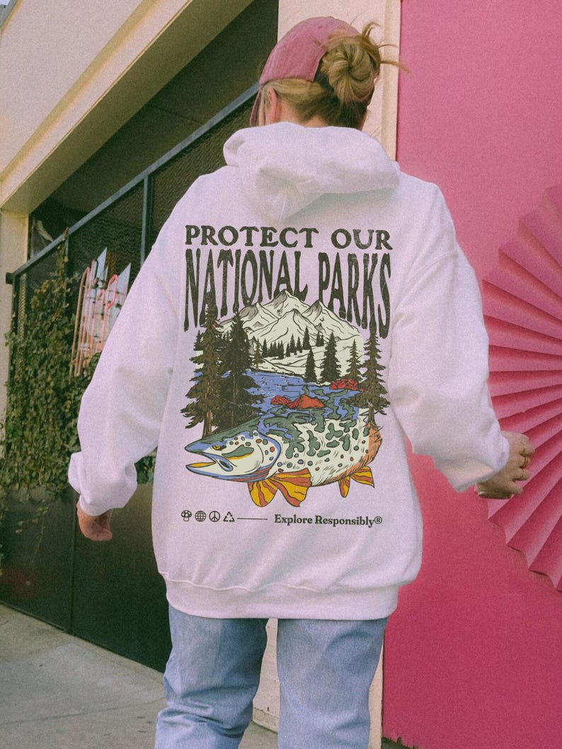 May include: A white hooded sweatshirt featuring a graphic on the back with the text PROTECT OUR NATIONAL PARKS and Explore Responsibly. The illustration depicts a mountain range, pine trees, and a large fish with colorful fins and scales.