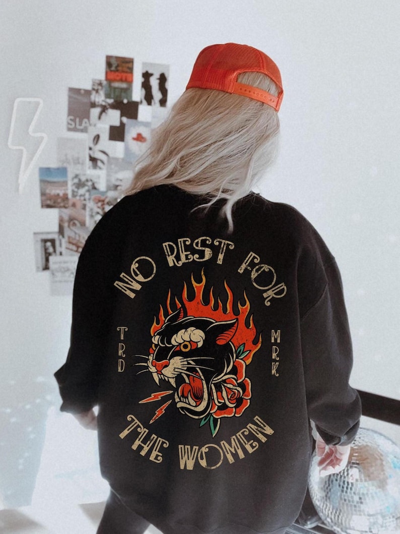 May include: Black sweatshirt with a graphic of a panther's head engulfed in flames, a red rose, and the text "NO REST FOR THE WOMEN." The sweatshirt is paired with an orange cap.