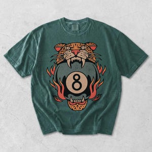 May include: Green t-shirt featuring a graphic of a tiger's head above a flaming eight ball. The tiger design is in shades of orange, brown, and black. The number eight is in a circle. The shirt is a casual wear item.