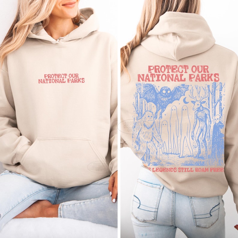 May include: A beige hooded sweatshirt featuring the red text "PROTECT OUR NATIONAL PARKS" on the front. The back displays a blue graphic of a forest with a bat, a yeti, ghosts, a deer-like creature, and an alien, with the text "PROTECT OUR NATIONAL PARKS" and "LEGENDS STILL ROAM FREE".