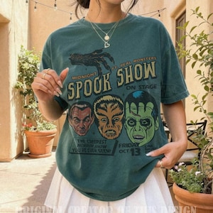 May include: A dark green short-sleeve t-shirt featuring a graphic print with a clawed hand and three monster faces. The text reads Midnight Spook Show, Ft. Real Monsters, On Stage, The Creepiest Horror Show You've Ever Seen, and Friday Oct. 13.