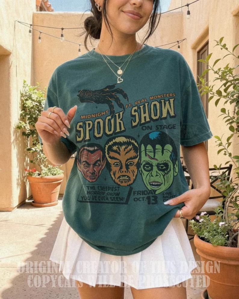 May include: A dark green short-sleeve t-shirt featuring a graphic print with a clawed hand and three monster faces. The text reads Midnight Spook Show, Ft. Real Monsters, On Stage, The Creepiest Horror Show You've Ever Seen, and Friday Oct. 13.