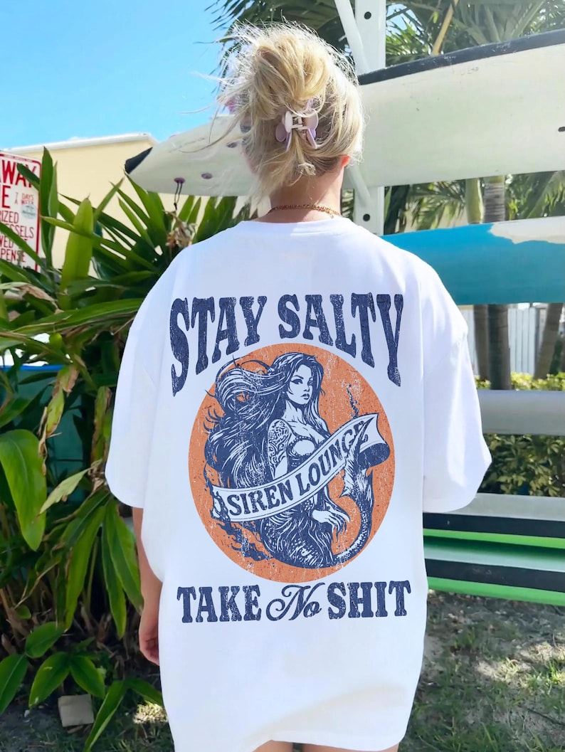 May include: White t-shirt with a graphic print of a mermaid with the text "Stay Salty Siren Lounge Take No Shit" on the back.