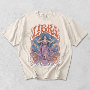 May include: A cream-colored short-sleeve t-shirt featuring a graphic print of a figure holding scales. The design includes the word LIBRA in large orange letters, a central purple-clad figure, blue sky elements, and orange floral accents.
