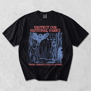 Peut inclure: Un t-shirt noir &agrave; manches courtes avec une impression graphique bleue repr&eacute;sentant une sc&egrave;ne de for&ecirc;t avec un hibou, un sasquatch, des fant&ocirc;mes et une cr&eacute;ature ressemblant &agrave; un cerf. Le texte indique PROTECT OUR NATIONAL PARKS et WHERE LEGENDS STILL ROAM FREE.