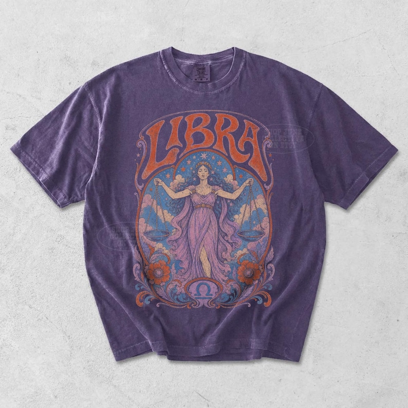 May include: A purple short-sleeve t-shirt featuring a graphic print of a figure holding scales, surrounded by floral and celestial motifs. The word LIBRA is printed in large orange letters at the top, with the Libra zodiac symbol at the bottom.