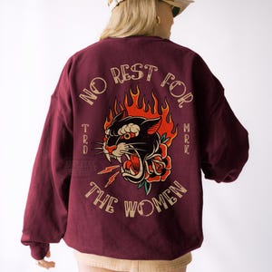 May include: Burgundy sweatshirt with a graphic on the back featuring a black panther with red flames, a rose, and the text "NO REST FOR THE WOMEN". The sweatshirt has long sleeves and a relaxed fit.