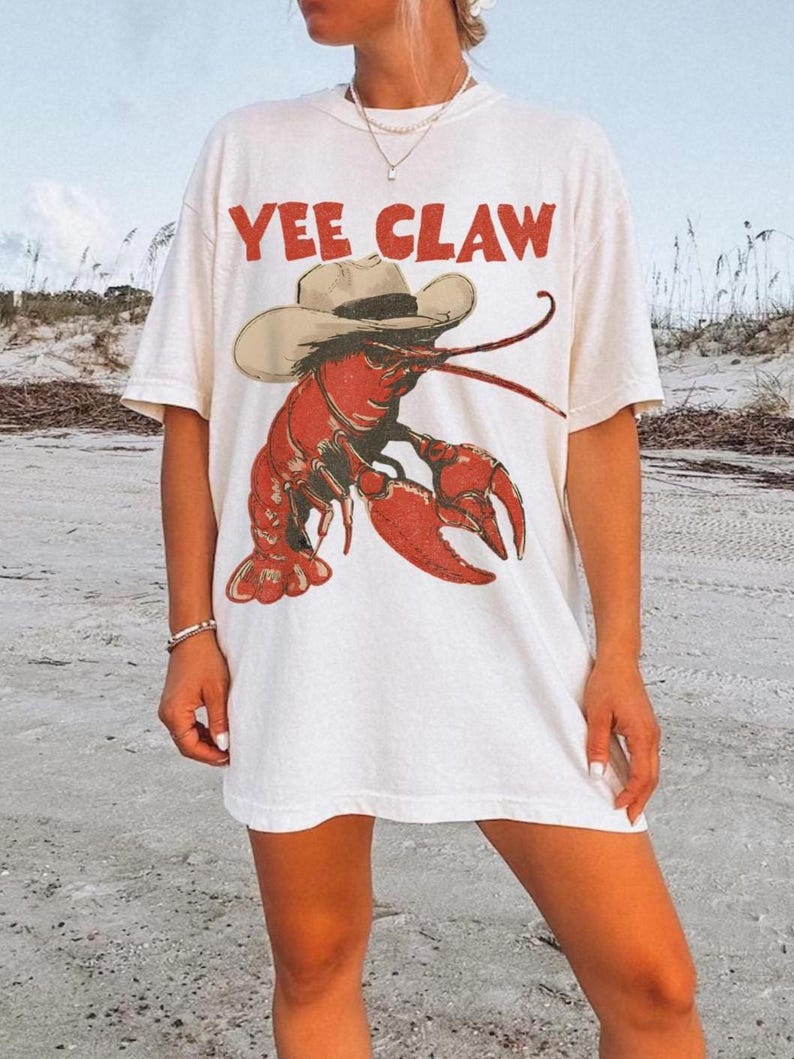Op de afbeelding: Wit T-shirt met een rode kreeft met een cowboyhoed. De tekst "YEE CLAW" is in het rood boven de kreeftafbeelding gedrukt. Het shirt heeft een relaxte, oversized pasvorm.