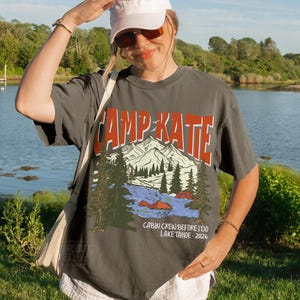 May include: A charcoal gray t-shirt with the text "CAMP KATE" in orange and white, featuring a mountain and lake graphic. The shirt also includes the text "CABIN CREW BEFORE I DO LAKE TAHOE - 2026".