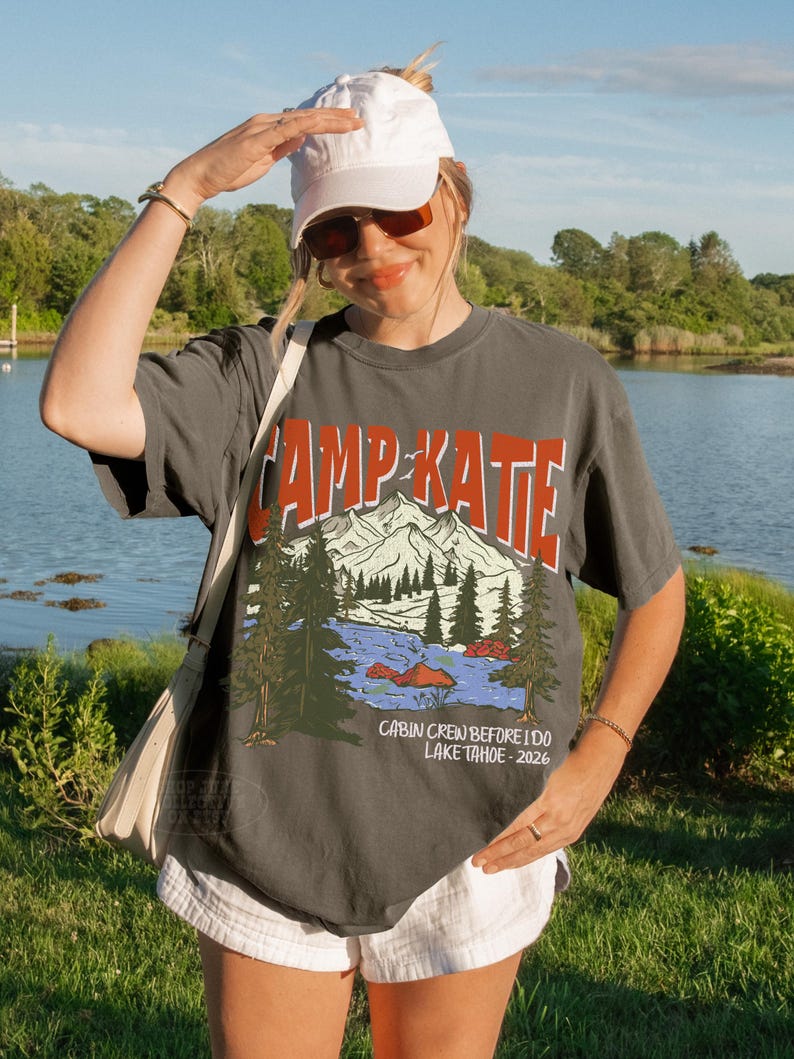 May include: A charcoal gray t-shirt with the text "CAMP KATE" in orange and white, featuring a mountain and lake graphic. The shirt also includes the text "CABIN CREW BEFORE I DO LAKE TAHOE - 2026".