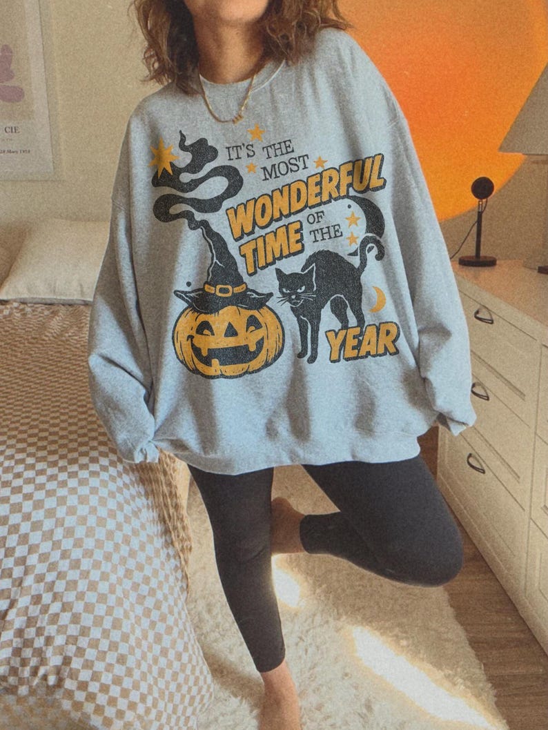 Peut inclure: Un sweat-shirt gris clair &agrave; col rond avec une impression graphique repr&eacute;sentant une citrouille d'Halloween portant un chapeau de sorci&egrave;re, un chat noir, des &eacute;toiles et un croissant de lune. Le texte indique "IT'S THE MOST WONDERFUL TIME OF THE YEAR".