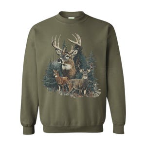 Puede incluir: Sudadera verde oliva con una ilustraci&oacute;n detallada de ciervos en un entorno forestal. El dise&ntilde;o incluye un gran ciervo con astas prominentes y dos ciervos m&aacute;s peque&ntilde;os. La obra de arte est&aacute; ambientada sobre un fondo de &aacute;rboles de hoja perenne.