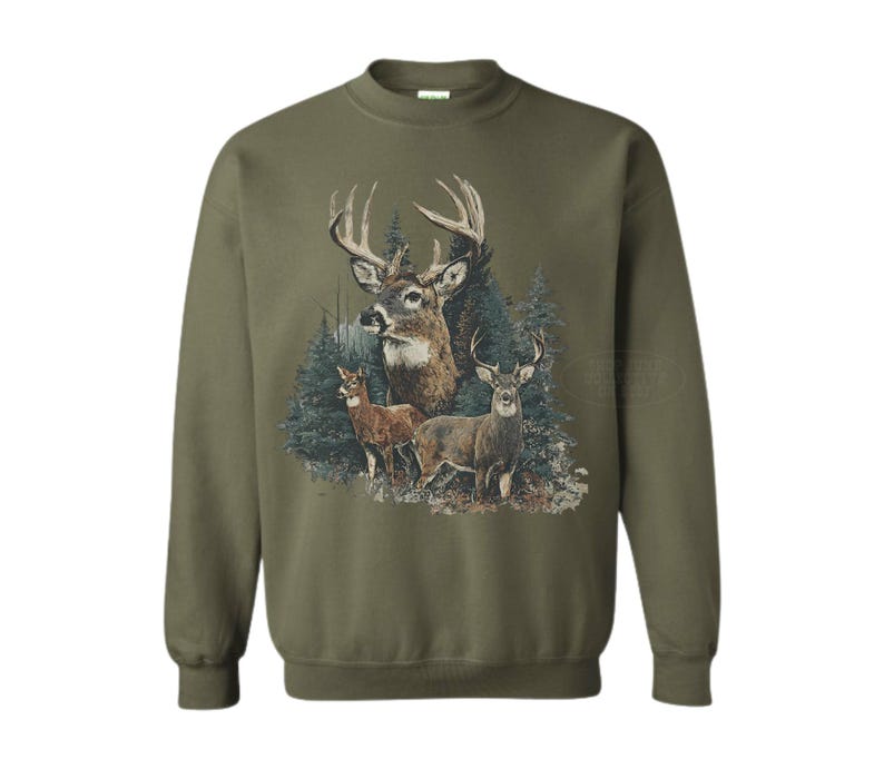 Puede incluir: Sudadera verde oliva con una ilustraci&oacute;n detallada de ciervos en un entorno forestal. El dise&ntilde;o incluye un gran ciervo con astas prominentes y dos ciervos m&aacute;s peque&ntilde;os. La obra de arte est&aacute; ambientada sobre un fondo de &aacute;rboles de hoja perenne.