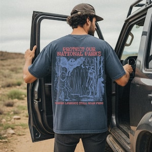 May include: A dark blue short-sleeve t-shirt featuring a graphic on the back with the red text "PROTECT OUR NATIONAL PARKS" and "WHERE LEGENDS STILL ROAM FREE". The illustration depicts a forest scene with a large owl, a deer, and a sasquatch.