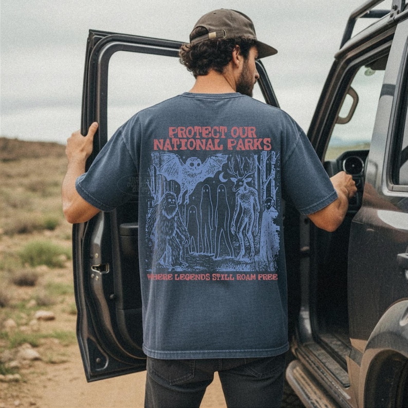 May include: A dark blue short-sleeve t-shirt featuring a graphic on the back with the red text "PROTECT OUR NATIONAL PARKS" and "WHERE LEGENDS STILL ROAM FREE". The illustration depicts a forest scene with a large owl, a deer, and a sasquatch.