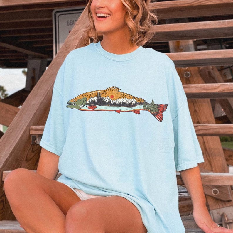 Puede incluir: Camiseta azul claro con un gr&aacute;fico detallado de un pez con un dise&ntilde;o de monta&ntilde;a y puesta de sol. El pez tiene un cuerpo marr&oacute;n, cabeza verde y cola roja. La camiseta es de material suave.