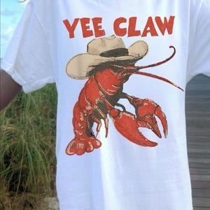 Op de afbeelding: Wit T-shirt met een rode afbeelding van een kreeft met een cowboyhoed. De tekst "YEE CLAW" staat boven de kreeft. Het shirt heeft korte mouwen.