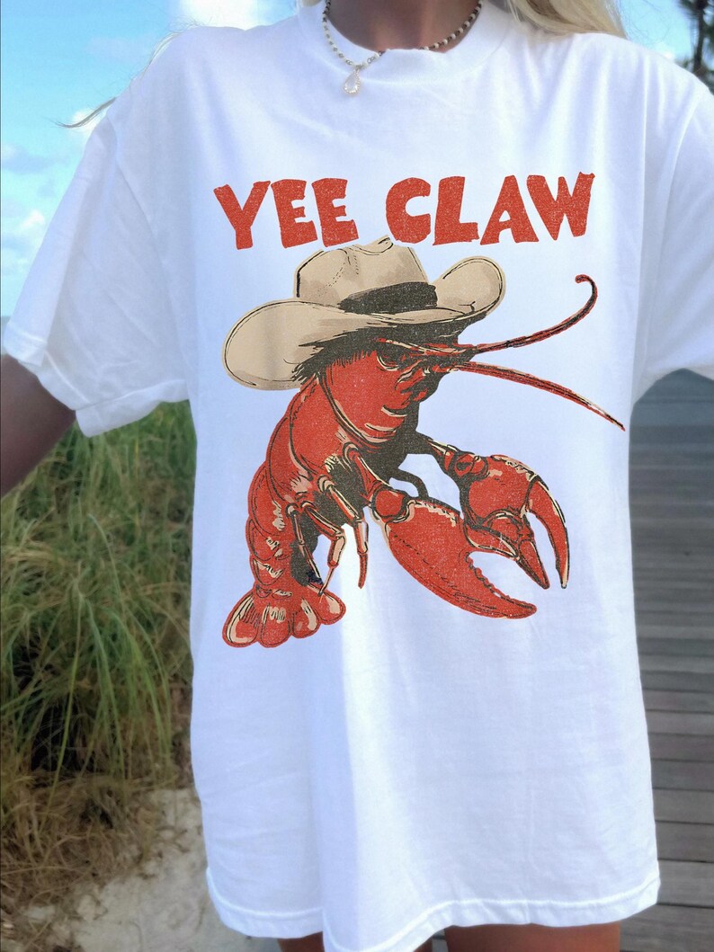 Op de afbeelding: Wit T-shirt met een rode afbeelding van een kreeft met een cowboyhoed. De tekst "YEE CLAW" staat boven de kreeft. Het shirt heeft korte mouwen.