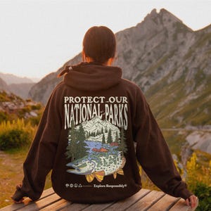 May include: A dark brown hooded sweatshirt featuring a graphic on the back with the text "PROTECT OUR NATIONAL PARKS" above an illustration of mountains, pine trees, and a large fish, with the text "Explore Responsibly" at the bottom.