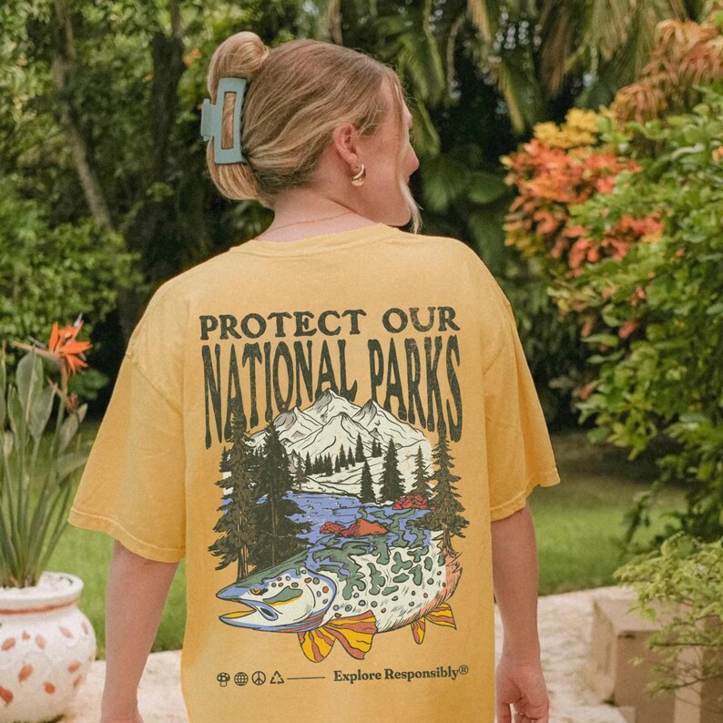 May include: A yellow t-shirt with a vintage-style graphic of a fish, mountains, and trees. The text "Protect Our National Parks" is printed in black on the shirt. The text "Explore Responsibly" is printed in black at the bottom of the shirt.