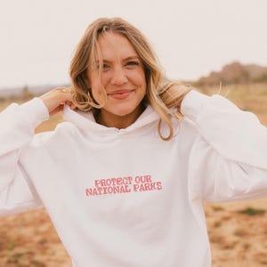 May include: A white long-sleeve hooded sweatshirt featuring the text "PROTECT OUR NATIONAL PARKS" printed in a distressed red font across the chest. The garment includes a hood with drawstrings and a front kangaroo pocket for storage.